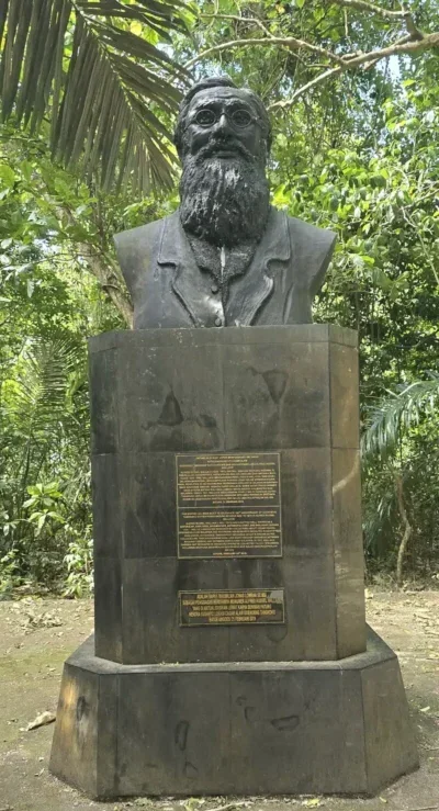 Busto de bronce de Alfred Russel Wallace sobre un pedestal de piedra en un entorno natural de Sulawesi.