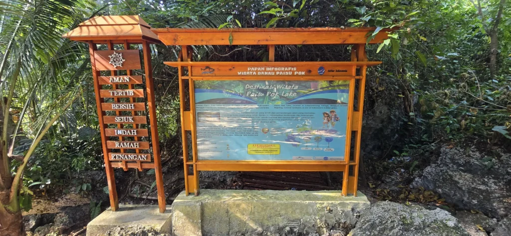 Cartel de madera con información turística y mapa en la entrada del Lago Paisupok, rodeado de vegetación tropical.