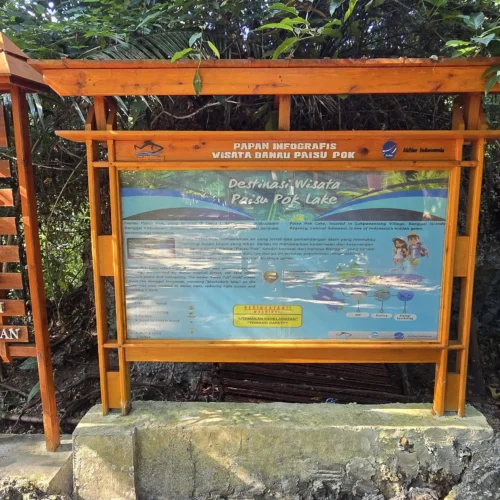 Cartel de madera con información turística y mapa en la entrada del Lago Paisupok, rodeado de vegetación tropical.