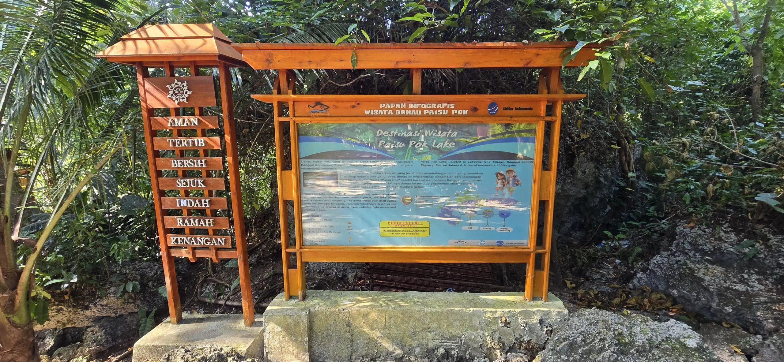 Cartel de madera con información turística y mapa en la entrada del Lago Paisupok, rodeado de vegetación tropical.
