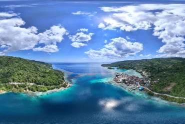 Vista panorámica aérea del pueblo de Paisupok y la Bahía Kawalu en las Islas Banggai, mostrando la costa, casas tradicionales, selva tropical y aguas turquesas.